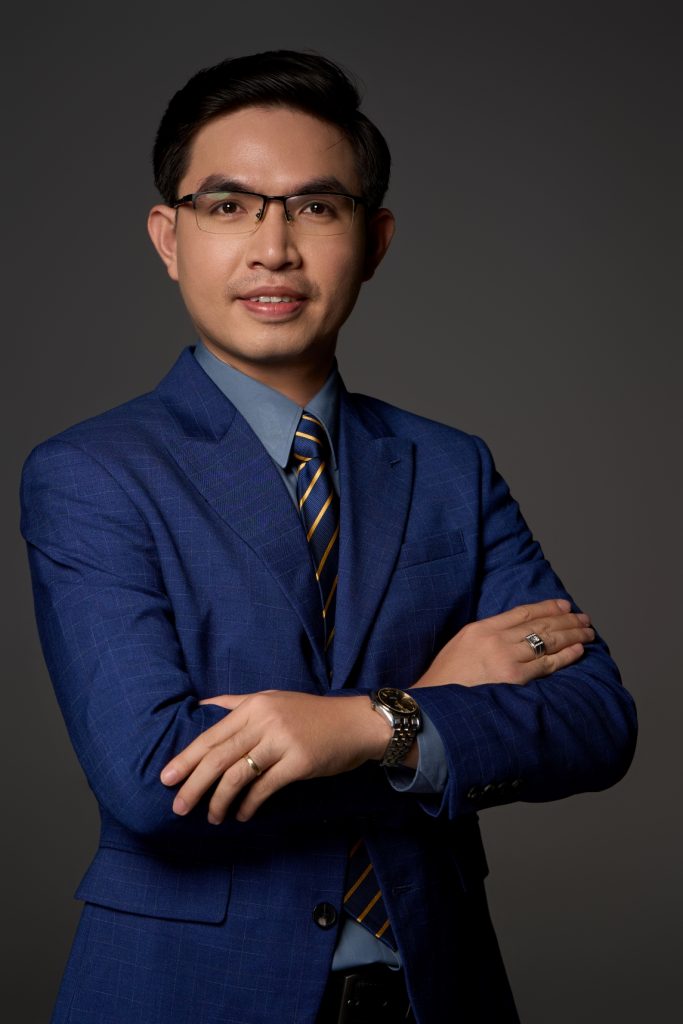 Chụp headshot doanh nhân nam đeo kính, mặc vest xanh navy thắt cà vạt sọc chéo, khung cận mặt trên phông nền xám tối, thực hiện bởi Lus Photo