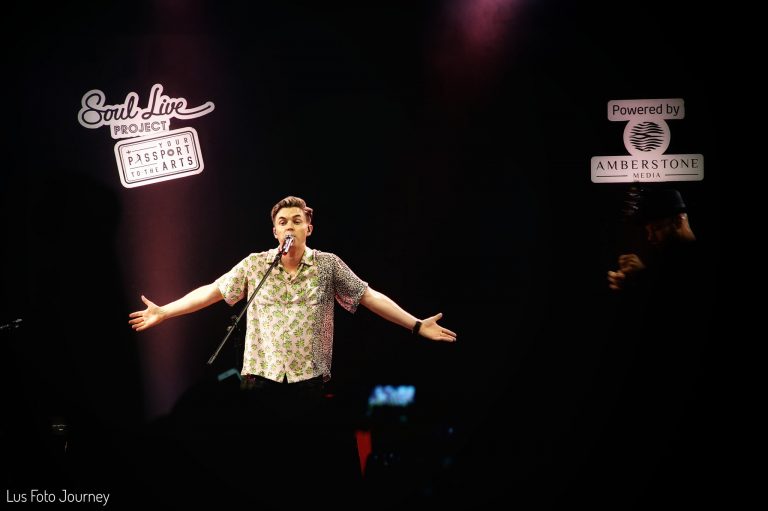 Chụp ảnh concert ca sĩ quốc tế: ca sĩ mặc áo sơ mi hoa dang hai tay trên sân khấu, bảng đèn neon phía sau, tông hồng tím, thực hiện bởi Lus Photo