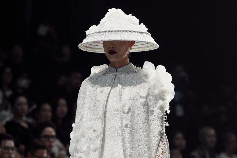 Chụp ảnh fashion show runway: người mẫu đội mũ vành lớn màu trắng, áo khoác ren trắng, bước trên sàn diễn nền tối, khán giả hai bên, thực hiện bởi Lus Photo