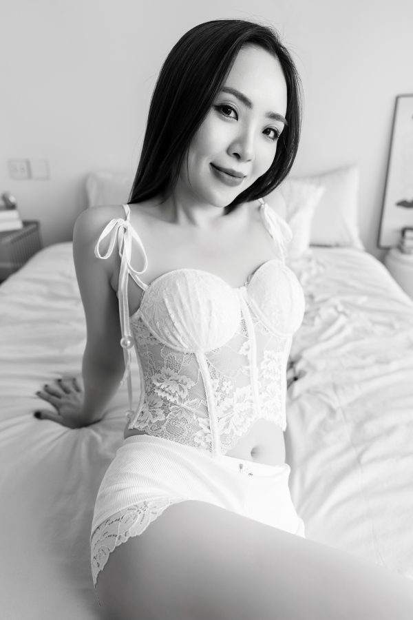 Ảnh boudoir trắng đen: cô gái ngồi trên giường, mặc corset ren trắng dây rút nơ vai, tóc dài xoã, không gian phòng ngủ tối giản, thực hiện bởi Lus Photo