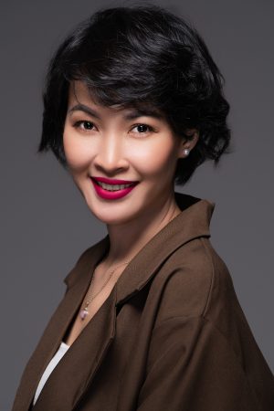 Chụp ảnh chân dung doanh nhân nữ tóc ngắn uốn xoăn mặc áo khoác nâu cổ bẻ to, trên phông nền xám, thực hiện bởi Lus Photo